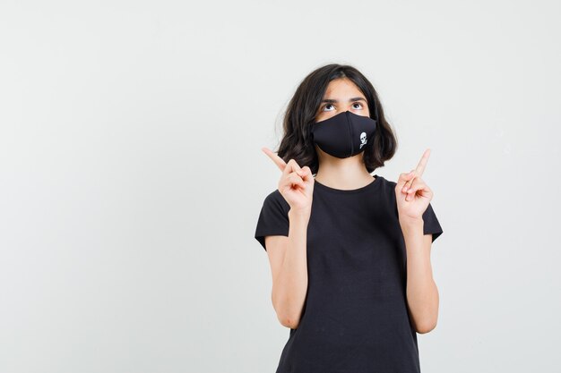 Klein meisje in zwart t-shirt, masker dat omhoog wijst en gericht, vooraanzicht kijkt.