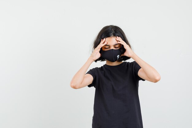 Klein meisje in zwart t-shirt, masker met sterke hoofdpijn en op zoek moe, vooraanzicht.