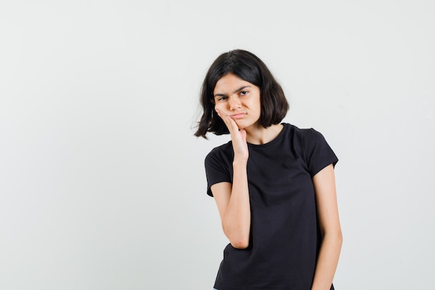 Klein meisje in zwart t-shirt met pijnlijke kiespijn en op zoek verdrietig, vooraanzicht.