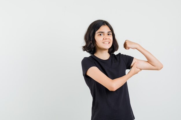 Klein meisje in zwart t-shirt wijzend op haar spieren en kijkt vrolijk, vooraanzicht.