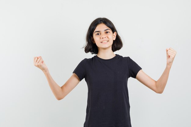 Klein meisje in zwart t-shirt winnaar gebaar tonen en op zoek zelfverzekerd, vooraanzicht.