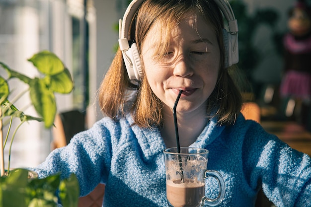 Klein meisje luistert naar muziek in een koptelefoon en drinkt cacao met zonnige ochtend
