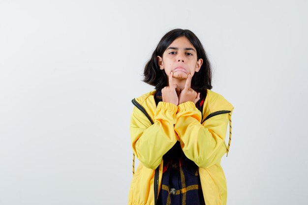 Klein meisje met droevig gebaar met vingers op gezicht in geruit overhemd, jasje en schattig, vooraanzicht.