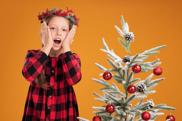 Klein meisje met een kerstkrans in een geruit hemd staat verbaasd naast een kerstboom over een oranje muur