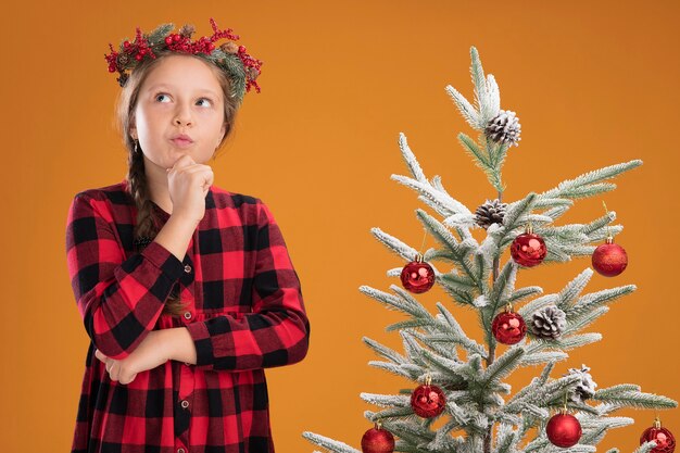 Klein meisje met een kerstkrans in een geruit overhemd en kijkt verbaasd op terwijl ze naast een kerstboom over een oranje muur staat