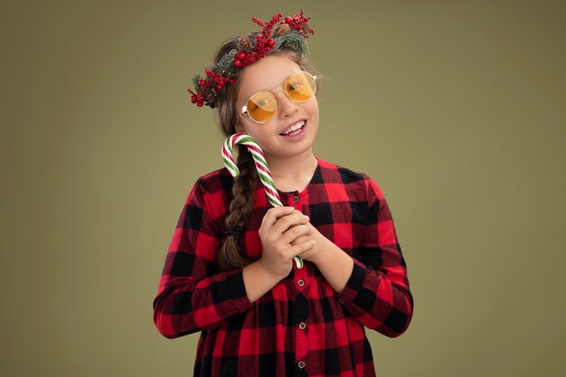 Gratis foto klein meisje met een kerstkrans in een geruite jurk met snoepgoed, blij en positief glimlachend, vrolijk over de groene muur