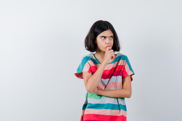 Klein meisje staat in denkende pose in t-shirt, spijkerbroek en kijkt peinzend. vooraanzicht.