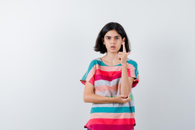 Klein meisje steekt wijsvinger op in eureka-gebaar terwijl ze de hand op de elleboog in t-shirt houdt en er verstandig uitziet. vooraanzicht.