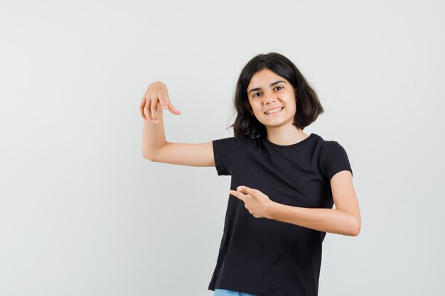 Klein meisje wijzend naar haar vinger in zwart t-shirt en kijkt vrolijk, vooraanzicht.