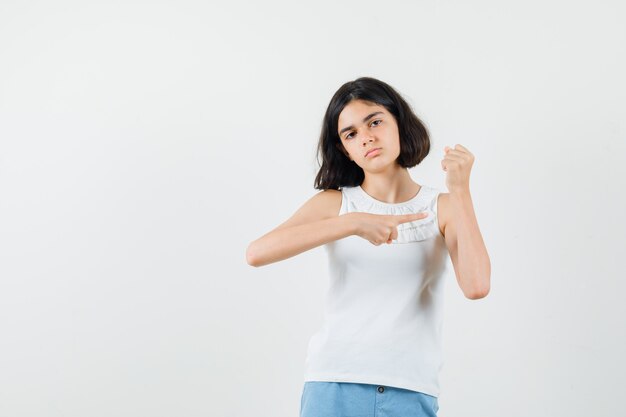 Klein meisje wijzend op haar opgeheven arm in witte blouse, korte broek en op zoek verdrietig, vooraanzicht.