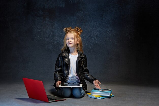 Klein meisje zit met laptop, tablet en telefoon in grijze studio