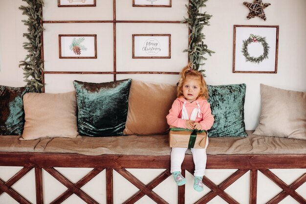 Klein mooi meisje met golvende haie zit van de bank met een doos met een cadeau