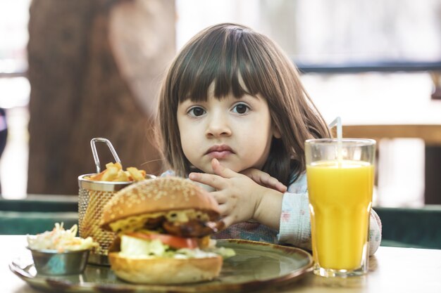 Klein schattig meisje een fastfood-sandwich met frietjes en jus d'orange eten in een café. Snel voedselconcept.