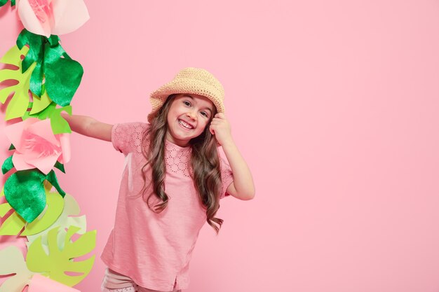 Klein schattig meisje in zomer hoed op kleur geïsoleerd roze achtergrond met papieren bloemen, plaats voor tekst, zomer reclame concept, Studio schieten