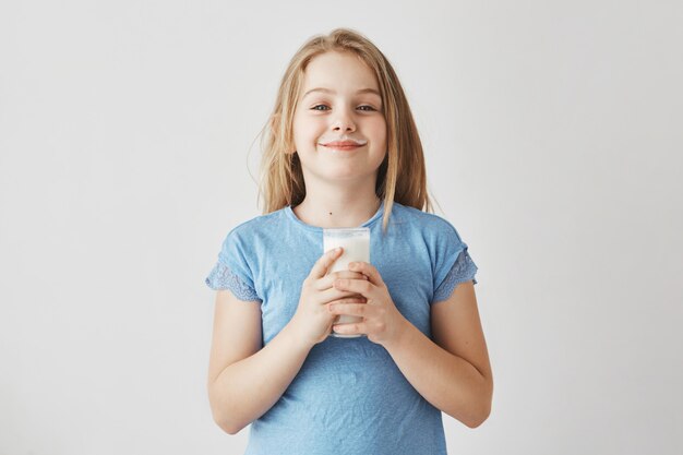 Klein schattig meisje met blond haar in blauw t-shirt met melk druppels op het gezicht, blij om haar dag te beginnen met een groot glas gezond drankje.