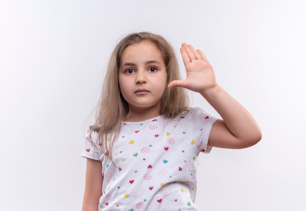 klein schoolmeisje dat wit t-shirt draagt dat hand op geïsoleerde witte muur opheft