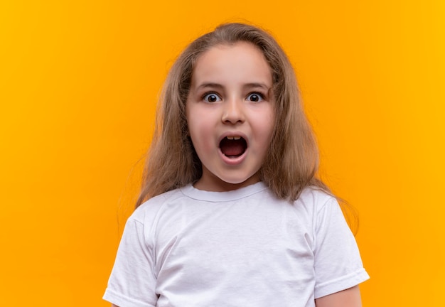 klein schoolmeisje dat wit t-shirt draagt dat mond op geïsoleerde oranje muur opent