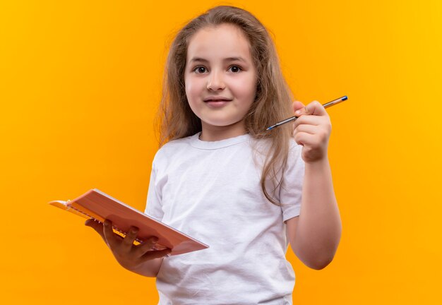 klein schoolmeisje die het witte notitieboekje en de pen van de t-shirtholding op geïsoleerde oranje muur dragen