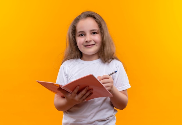 Klein schoolmeisje die het witte notitieboekje en de pen van de t-shirtholding op geïsoleerde oranje muur dragen