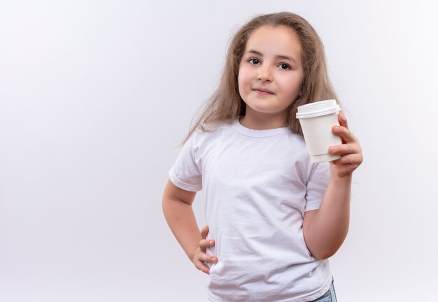 klein schoolmeisje die wit t-shirt dragen dat kopje koffie houdt, legde haar hand op de heup op afgelegen witte muur