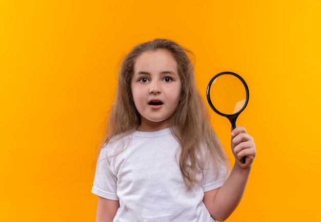 klein schoolmeisje die wit t-shirt dragen die vergrootglas op geïsoleerde oranje muur houden