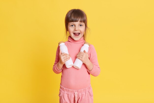 Klein schoonheidsmeisje die in roze kleren met bruin haar twee flessen melk op geel houden