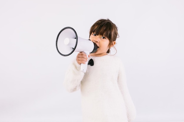 Klein zwartharig meisje dat een witte trui met vlinderdas draagt en door een witte megafoon spreekt.