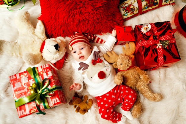 Kleine baby in rode en witte kleren ligt tussen kerstcadeautjes en speelgoed op de vloer