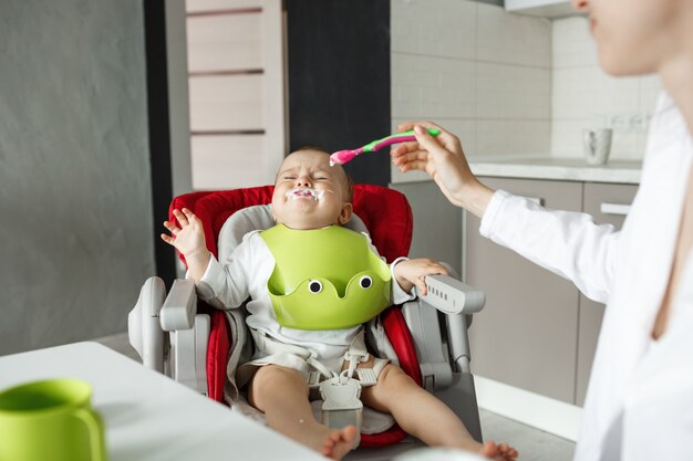 Kleine baby zittend in een kinderstoel met restjes in de buurt van de mond huilt en weigert te eten terwijl moeder hem probeert te voeden met een lepel.