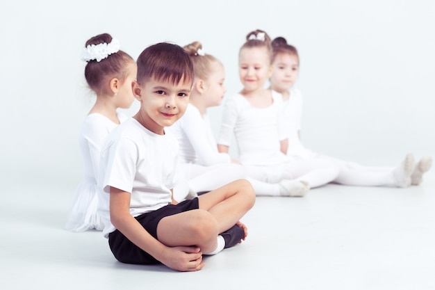 Kleine ballerina's en jongen ballerun doen oefeningen en zitten op de vloer in witte balletles.
