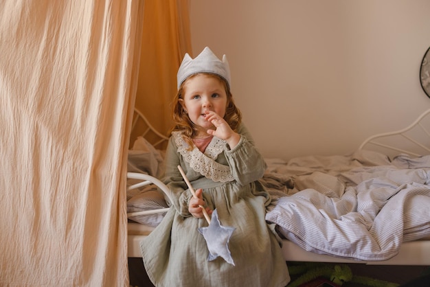 Kleine blanke roodharige baby in jurk speelt prinses terwijl hij op bed zit in de kinderkamer peuterconcept