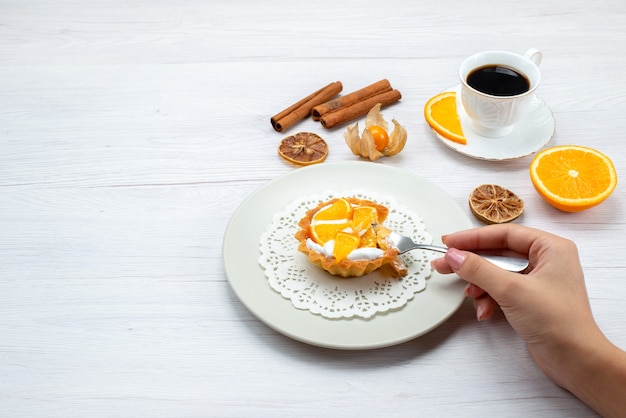 kleine cake met room en gesneden sinaasappelen die door vrouw worden gegeten, samen met koffie en kaneel op licht bureau, fruitcake koekje zoete suiker