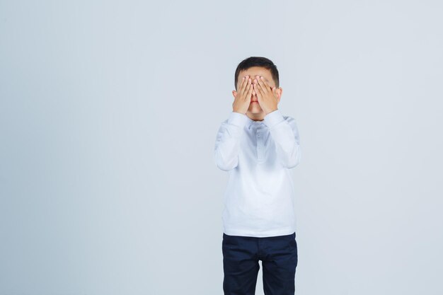 Kleine jongen in wit overhemd, broek die handen op het gezicht houdt en opgewonden kijkt, vooraanzicht.