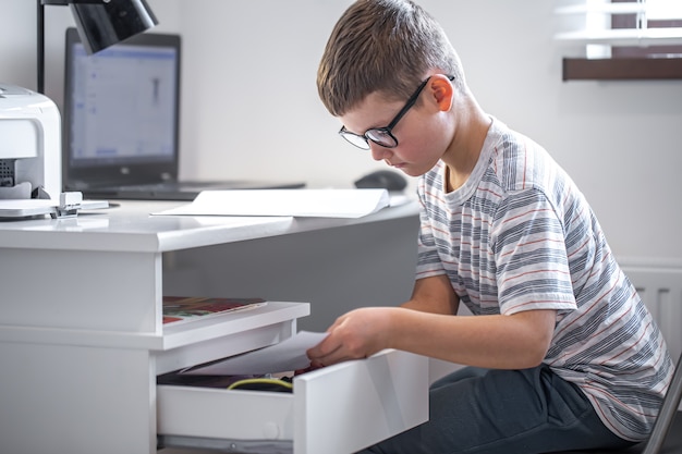 Kleine jongen met bril zit aan zijn bureau voor een laptop op zoek naar iets in een la.