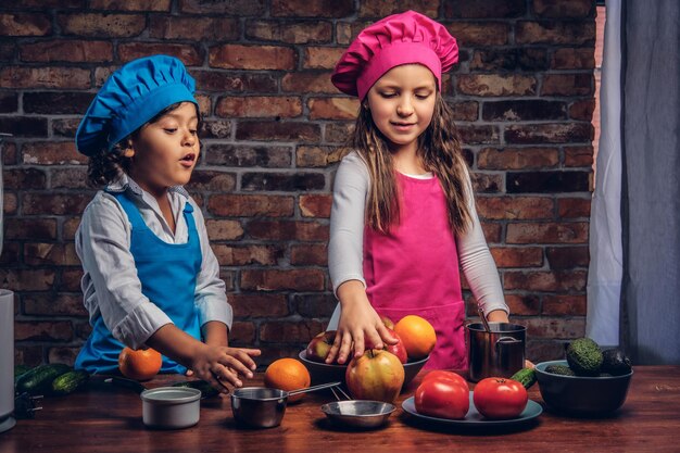 Kleine jongen met bruin krullend haar gekleed in een blauwe kok uniform en een mooi meisje gekleed in een roze kok uniform samen koken in een keuken tegen een bakstenen muur. Leuk klein kokspaar.