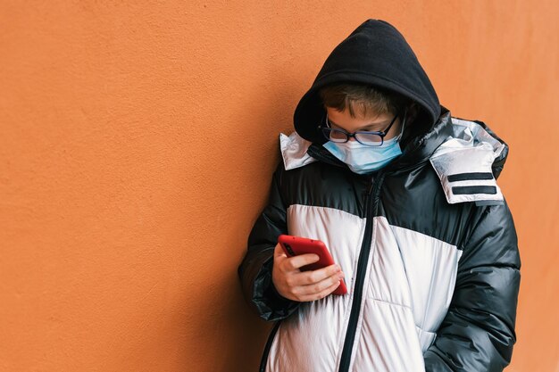 Kleine jongen met een hygiënisch masker, bril en winterkleding met zijn telefoon