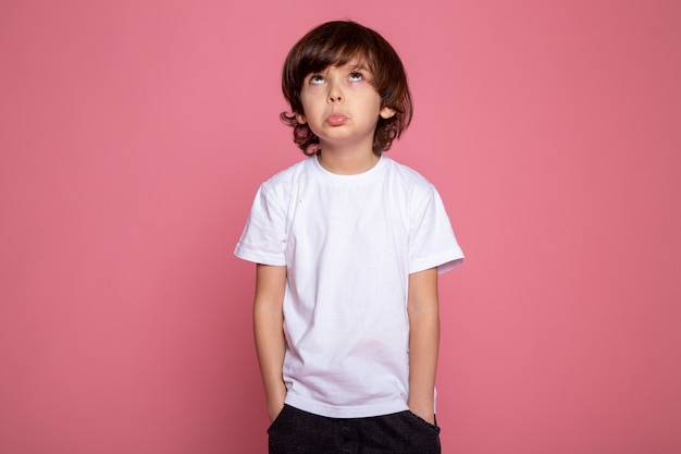 Kleine jongen met handen in zijn zak wit t-shirt en spijkerbroek kijken naar de celing op roze bureau