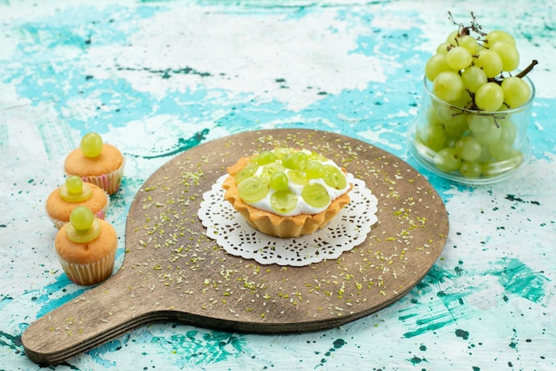 kleine lekkere cake met heerlijke room en gesneden en verse druivenkoekjes op blauw lichtbureau, fruit van de cake het zoete suiker