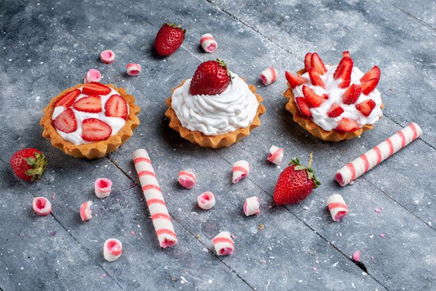 kleine romige cakes met gesneden en verse aardbeien samen met stoksnoepjes op grijs bureau, zoete kleur van fruitcake