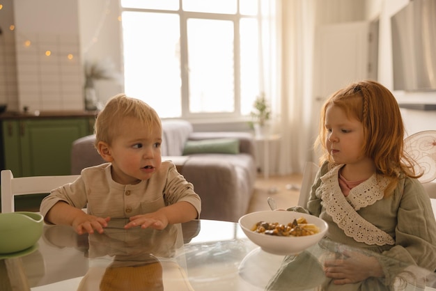 Kleine schattige blanke blonde jongen kijkt naar zijn oudere roodharige zus aan de ontbijttafel babyvoeding concept