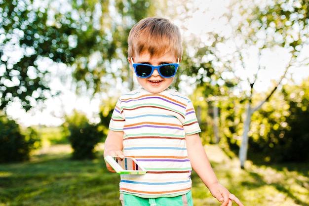Kleine schattige jongen telefoon bedrijf in zijn hand en glimlacht in de zomertuin.