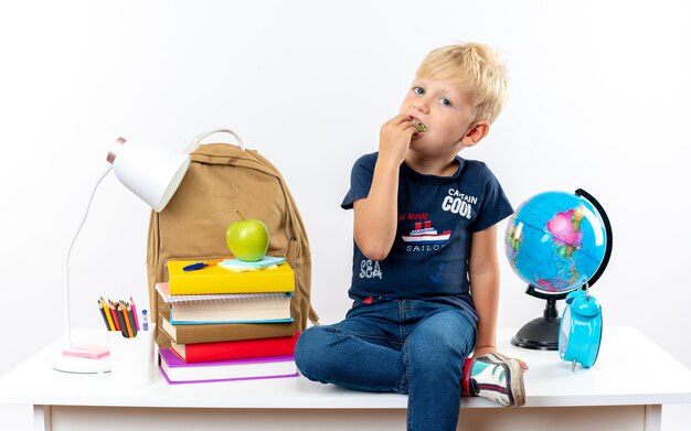 kleine schooljongen zittend op de tafel met schoolhulpmiddelen die een stuk appels eten