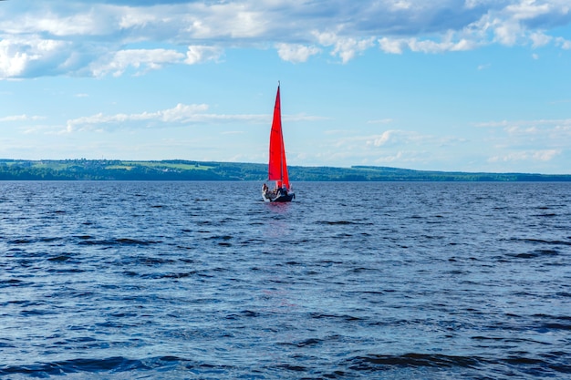 Kleine zeilboot met rode zeilen in het midden van een brede watervlakte met een beboste kust