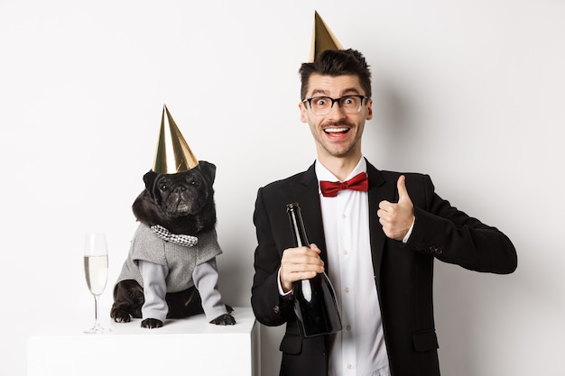 Kleine zwarte hond met feestmuts en staande in de buurt van gelukkige man die vakantie viert, eigenaar die duim omhoog laat zien en champagnefles vasthoudt, witte achtergrond