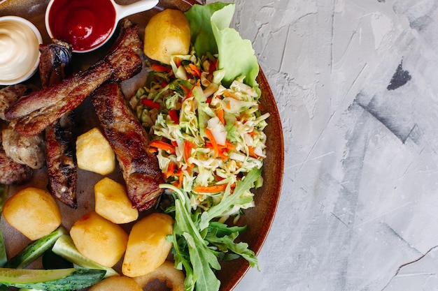 Kleiplaat vol bierhapjes staande op witte tafel Groentesalade gekookte aardappelen gegrilde kippenpoten geroosterde gouden ui en worstjes komkommers tomaten en sauzen ketchup en mayo