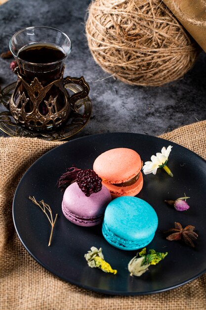 Kleurrijke macarons in een zwarte schotel en een glas thee op een rustieke jute.