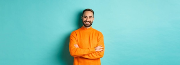 Gratis foto knap bebaard mannelijk model in oranje trui die er zelfverzekerd uitziet en glimlachende gekruiste armen op de borst stan