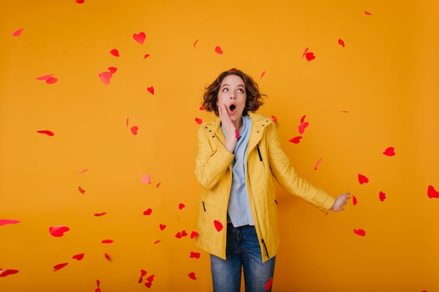 Knap bleek meisje in gele outfit die vliegende harten bekijkt. Studio shot van verbazingwekkende krullende vrouw in casual kleding met plezier in Valentijnsdag.