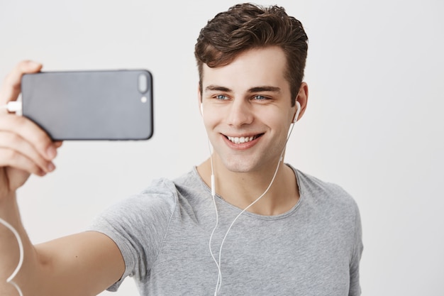 Knap kaukasisch jong mannetje met donker haar en aantrekkelijke blauwe ogen die mobiele telefoon houden, stellend voor selfie, kijkend met flirterige glimlach. knappe kerel die videogesprek voert.