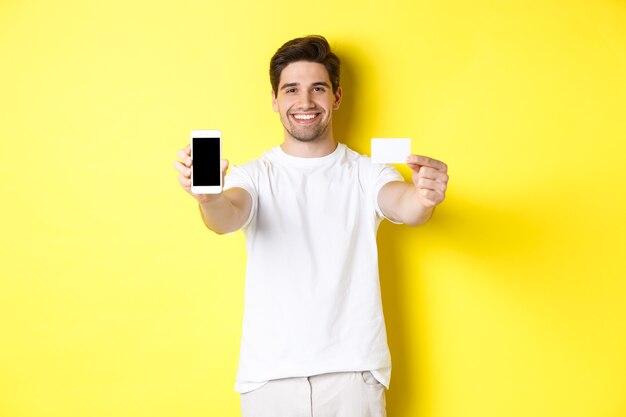 Knap kaukasisch mannelijk model dat het smartphonescherm en creditcard, concept mobiel bankieren en online winkelen, gele achtergrond toont.